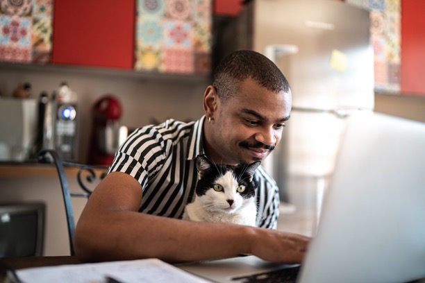 A photograph of a man in a black and white striped shirt with a black and white cat in his lap. The man is using a computer.